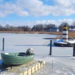 Ein grünes Boot liegt auf einer Eisfläche neben einem Leuchtturm. Im Hintergrund sind Häuser und Bäume zu sehen.