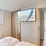 Schlafzimmer mit Bett, TV und Fenster. Durchgang zu Wohnbereich mit Tisch und Stühlen.