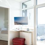 Living area with TV, red ottoman, and balcony view. Bedroom in background.