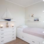 Bedroom with white bed and dresser, wall shelves with decor and model sailboat.