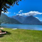 A quiet lakeside area with sun loungers under a tree and mountains in the background.