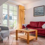 Ein Wohnzimmer mit rotem Sofa, grauem Sessel und Holztisch. Durchgehendes Fenster mit orangefarbenen Vorhängen.