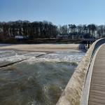 Holzbrücke führt zum Strand. Im Hintergrund sind Bäume und Gebäude zu sehen.