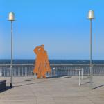Holzplattform mit Metallgeländer, Skulptur und Blick auf Meer unter blauem Himmel.