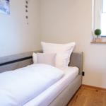 Ein Schlafzimmer mit Bett, Fenster und Schreibtisch. Wandbild und Pflanzen sind sichtbar.