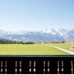 Blick von Balkon auf grünes Feld, Weg und schneebedeckte Berge.