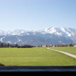Weitläufiges Grünfeld mit Pfad, dahinter Berglandschaft mit Schneekappen und Siedlung.