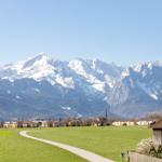Weitläufiges Feld mit Pfad, Dorf und schneebedeckten Bergen im Hintergrund.