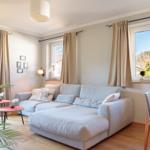 Wohnzimmer mit Sofa, Tisch und Blick auf Berge. Holzboden und Fenster mit Vorhängen.
