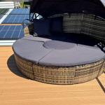 Round rattan lounge area with dark cushions and sunshade on terrace.