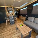 Living area with sofa, dining table, and kitchenette. Wooden floor and windows with blinds.