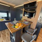 Kitchen area with wooden counter, stools, and provisions. Background: TV and sink.