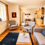 Wohnzimmer mit Sofa, zwei Sesseln, Tisch und Teppich. Küche und Essbereich im Hintergrund.
