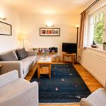Wohnzimmer mit Sofa, Sesseln, Teppich und TV. Fenster mit Blick auf Grün.