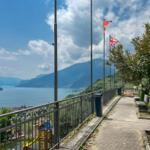 Weg mit Geländer und Flaggen überblickt den See und Berge.