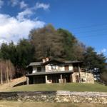 Haus mit Steinmauer und Terrasse auf grünem Hang unter blauem Himmel.
