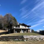 Haus auf grüner Anhöhe mit Steinmauer und Wald im Hintergrund unter blauem Himmel.