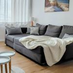 Ein grauer Sofa mit Kissen und einer Decke. Zwei runde Tische stehen auf dem Holzboden.