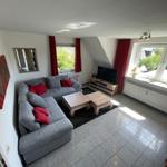 Wohnzimmer mit grauem Sofa, roten Kissen, Holztischen und großen Fenstern.