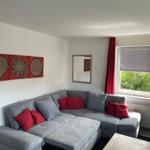 Ein Wohnzimmer mit grauem Ecksofa, roten Kissen und einem Holztisch. An der Wand hängt ein Bild.