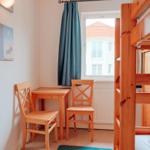 Zimmer mit Holzbett, Tisch und zwei Stühlen. Fenster mit Vorhang und Wandbild.