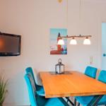 Esszimmer mit Holztisch, blauen Stühlen und Wandbild. An der Wand hängt ein Fernseher.