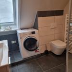 Bathroom with washing machine, toilet, and shower. Window with plant.