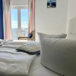 Ein Bett mit weißen Laken und Kissen. Im Hintergrund ein Fenster mit Blick auf Balkon und Meer.