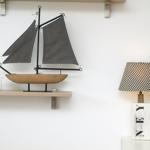 Ein Modellboot mit grauen Segeln steht auf einer Holzregal. Daneben steht eine Lampe mit kariertem Schirm.