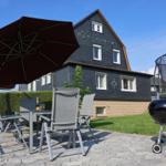 Terrasse mit Tisch, Stühlen, Sonnenschirm und Grill. Hintergrund: Haus mit Schieferdach.