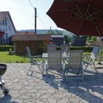 Terrasse mit Tisch, Stühlen, Sonnenschirm und Grill. Hintergrund: Haus und Landschaft.