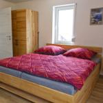 Schlafzimmer mit Holzbett, Kleiderschrank und Fenster. Nebenraum mit Waschmaschine und Spiegel.