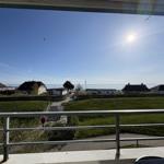 Blick vom Balkon auf Grünfläche, Häuser und Meer unter blauem Himmel mit Sonne und Möwen.