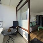 Balcony with table and chair overlooks green landscape. Living room with sofa, green cushions, and wooden floor.