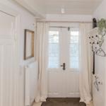 Hausflur mit weißen Wänden, Tür und Gardinen. An der Wand hängt ein Spiegel und ein Kleiderständer.