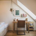 Ein Schlafzimmer mit Schreibtisch, Bett und Dachschräge. An der Wand hängt ein Bild und eine Pflanze.