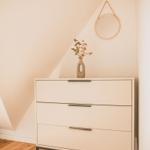 Schrank mit drei Schubladen, Vasen mit Blumen und runder Wandspiegel.