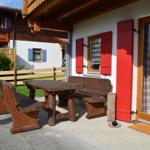 Terrasse mit Holztisch und Bänken, rote Fensterläden, Glastür mit kariertem Vorhang.