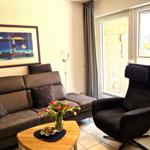 Ein Wohnzimmer mit Sofa, Sessel, Tisch und Blumen. An der Wand hängt ein Bild.