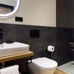 Badezimmer mit Waschbecken, Toilette und Dusche. Schwarze Wandfliesen und runder Spiegel.