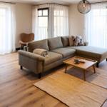 Wohnbereich mit Sofa, Tisch und Fenstern. Holzboden und helle Vorhänge.