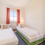Zwei Einzelbetten mit weißen Bettwäschen und Kissen. Fenster mit roten Vorhängen und Wandbild.