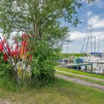 Ruderboote und rote Flaggen stehen unter einem Baum am Bootssteg. Segelboote liegen im Hafen.