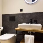 Badezimmer mit Toilette, Waschbecken und Spiegel. Schwarze Wandverkleidung und Holzunterschrank.