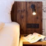Bettkopf aus Holz mit Wandlampe und Steckdose. Auf Nachttisch liegt eine Zeitschrift.