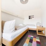 Zimmer mit zwei Betten, Holzmöbeln und buntem Teppich auf dunklem Holzfußboden.