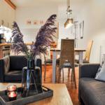 Wohnzimmer mit Esstisch, Sofa und Küchenbereich. Holztisch, Sessel und Vase mit Pflanzen.