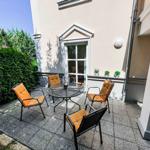 Terrasse mit Tisch und vier Stühlen, orangefarbene Sitzkissen, Pflanzen und Fenster im Hintergrund.