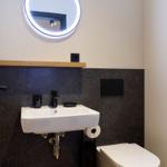 Badezimmer mit Waschbecken, Toilette und runden Spiegel mit Beleuchtung.