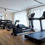 Fitnessraum mit Laufband, Ellipsentrainer und Gewichten. Holzfußboden und große Fenster.